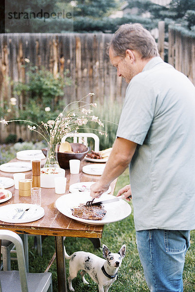 Ein Mann  der bei einer Familienmahlzeit in einem Garten Fleisch tranchiert und dabei von einem kleinen Hund unter dem Tisch beobachtet wird.