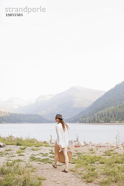 Eine Frau mit Hut und weißem Hemd mit einem Korb  am Ufer eines Bergsees.