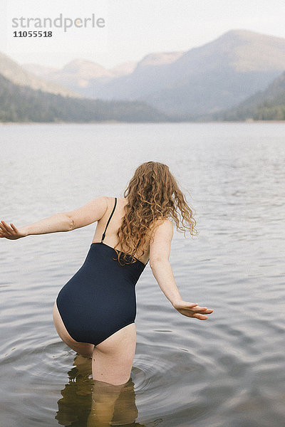 Eine Frau in einem schwarzen Badeanzug  die in einen ruhigen See in den Bergen watet.