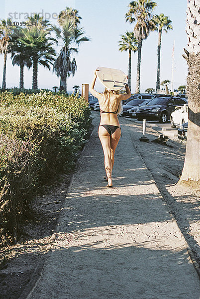 Eine Frau in einem schwarzen Bikini  die mit einem Surfbrett auf dem Kopf einen Weg entlang läuft.