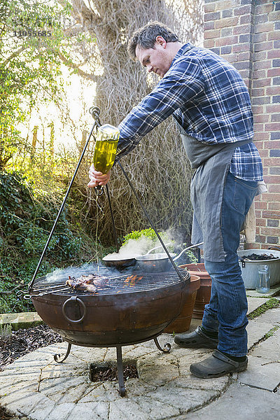 Ein Mann kocht Wildvögel über offenem Feuer und gießt Olivenöl aus einer Flasche auf das Fleisch.