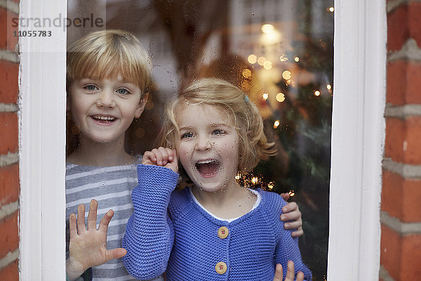 Zwei Kinder  ein Junge und ein Mädchen  blicken mit aufgeregten Gesichtsausdrücken aus einem Fenster zu Hause.