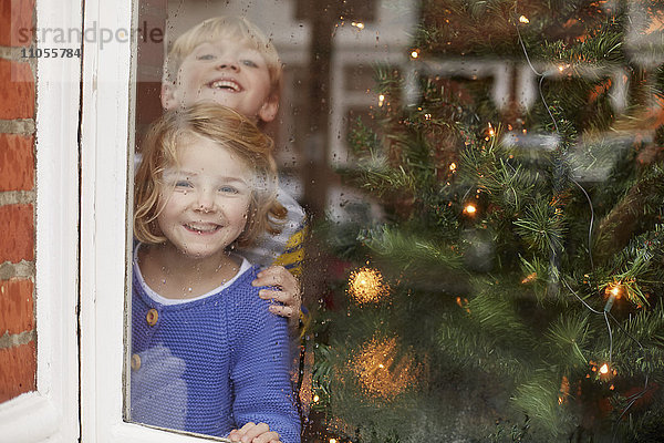 Zwei Kinder  ein Junge und ein Mädchen  schauen zu Hause neben einem Weihnachtsbaum aus dem Fenster.