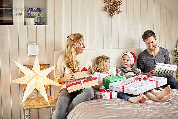 Eine Familie  zwei Erwachsene und zwei Kinder sitzen am Weihnachtsmorgen im Bett und öffnen gemeinsam Geschenke.