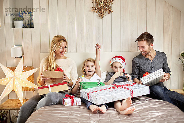 Eine Familie  zwei Erwachsene und zwei Kinder sitzen am Weihnachtsmorgen im Bett und öffnen gemeinsam Geschenke.
