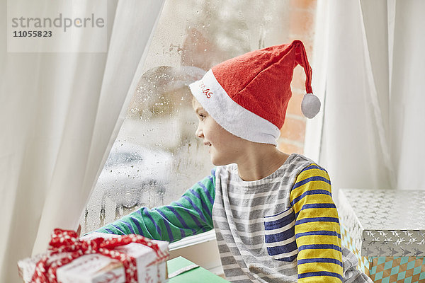 Weihnachtsmorgen in einem Familienhaus. Ein Junge mit Weihnachtsmannmütze  der aus einem Schlafzimmerfenster schaut.