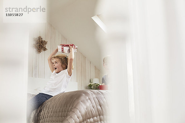 Weihnachtsmorgen in einem Familienhaus. Ein Mädchen hält ein Geschenk über ihrem Kopf.