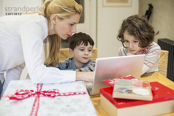 Zwei Kinder und ihre Mutter schauen auf einen Laptop-Computerbildschirm um einen Küchentisch herum.