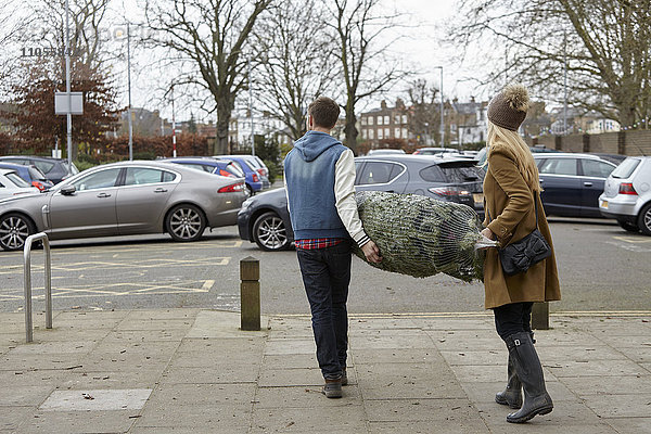 Zwei Personen  die einen netzartigen Weihnachtsbaum zum Parkplatz tragen.