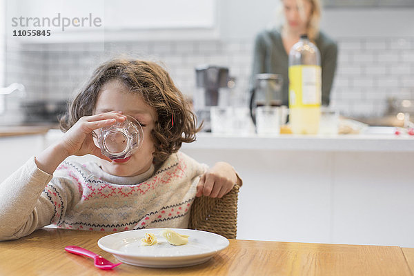 Ein Kind  das an einem Tisch sitzt und aus einem Glas trinkt  eine Frau  die hinter ihr in einer häuslichen Küche steht.
