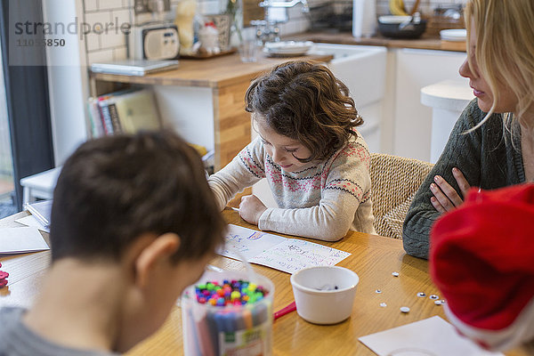 Drei Kinder und eine erwachsene Frau an einem Tisch  malen und schreiben Karten und Briefe an den Weihnachtsmann.