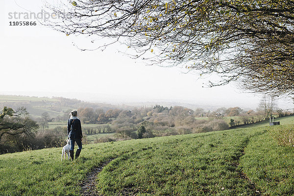 Eine Frau geht mit einem Hund auf einer Anhöhe mit Blick auf die Landschaft spazieren.