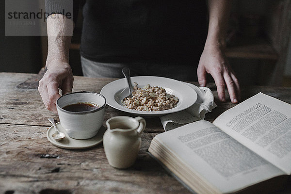 Eine Person an einem Tisch mit einer Tasse Kaffee  einer Schüssel Müsli und einem aufgeschlagenen Buch.