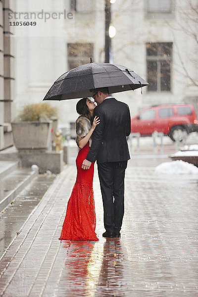 Eine Frau in einem langen roten Abendkleid mit Fischschwanzrock und Pelzstola und ein Mann im Anzug  der sich auf einer Straße unter einem Regenschirm küsst.