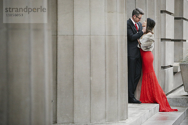 Eine Frau in einem langen roten Abendkleid mit Fischschwanzrock und Pelzstola und ein Mann in einem Anzug  die sich auf den Stufen eines Gebäudes umarmen.