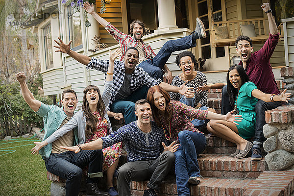 Eine Gruppe von Freunden sitzt auf den Stufen einer Hausveranda  posiert und lacht  die Arme ausgestreckt.