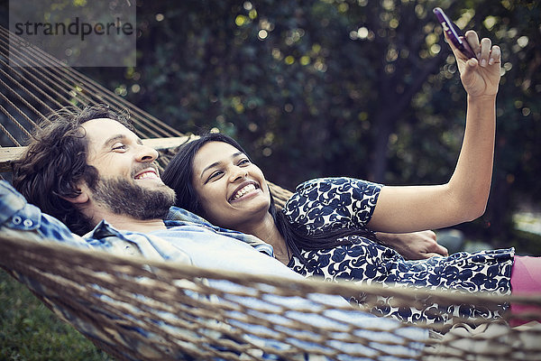 Ein Ehepaar  ein junger Mann und eine junge Frau  die in einer großen Hängematte im Garten liegen und sich ein Selfy von sich selbst nehmen.