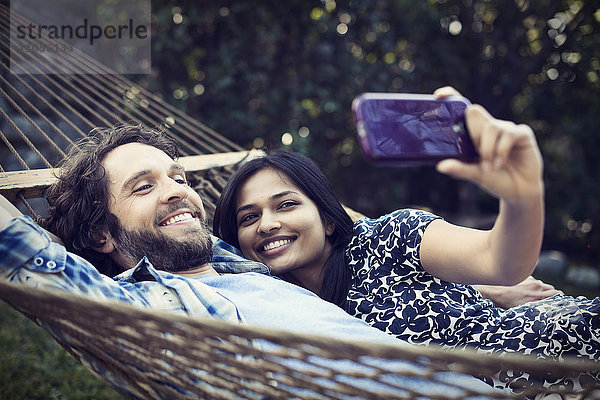 Ein Ehepaar  ein junger Mann und eine junge Frau  die in einer großen Hängematte im Garten liegen und sich ein Selfy von sich selbst nehmen.