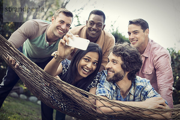 Eine Gruppe von Freunden  die sich in einer großen Hängematte im Garten bei einem Bier und einem Selfie ausruhen.