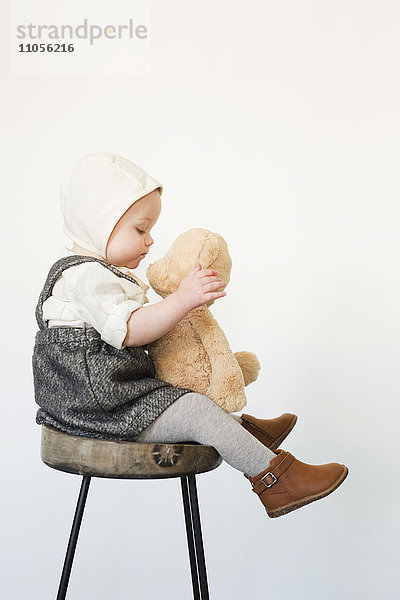 Ein kleines Kind  ein Mädchen  das auf einem hohen Hocker sitzt und einen Teddybär hält.
