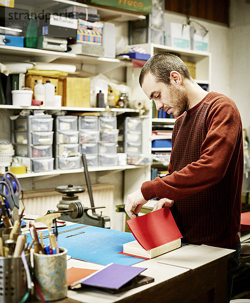 Ein Mann  der in einer Buchbinderei arbeitet und einen roten Einband für frisch geheftete Seiten herstellt.
