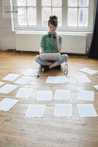 Junge Frau  die in einem Probenstudio mit einem Laptop auf dem Boden sitzt und sich Notenblätter ansieht.