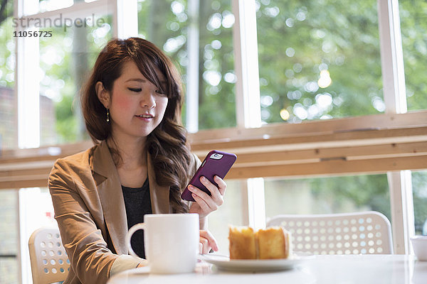 Lächelnde junge Frau  die mit einem Becher und einem Stück Kuchen an einem Tisch sitzt und auf ihr Smartphone schaut.