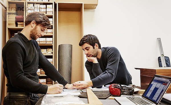 Eine Möbelwerkstatt  die maßgeschneiderte zeitgenössische Möbelstücke unter Verwendung traditioneller Fertigkeiten im modernen Design herstellt. Zwei Personen diskutieren über einen Entwurf und beziehen sich dabei auf Zeichnungen und Laptop-Computer.