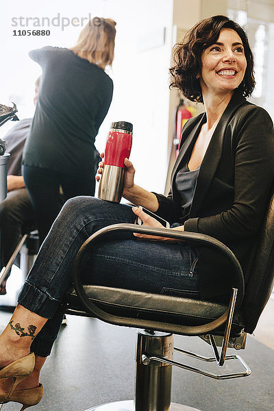 Eine Kundin sitzt in einem Friseursalonstuhl und hält eine Tasse Kaffee in der Hand.