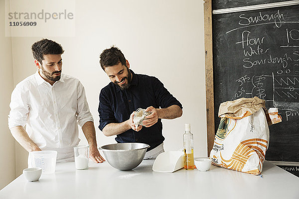 Zwei Bäcker stehen an einem Tisch und bereiten Brotteig  Backzutaten und eine Tafel an der Wand vor.