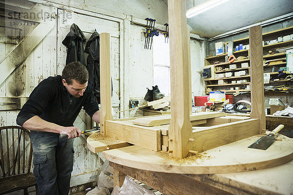 Ein Mann in einer Schreinerei  der mit einem Hammer an der Kante eines neuen Holztisches arbeitet.