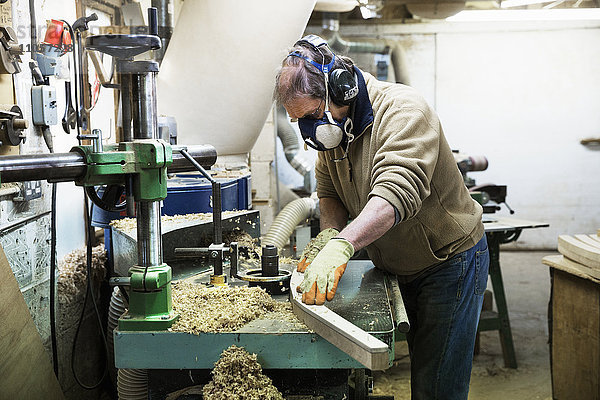 Mann steht in einer Schreinerei  trägt ein Atemschutzgerät und Gehörschutz und arbeitet an einem Stück Holz.