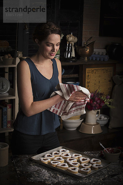 Valentinstag beim Backen  junge Frau steht in einer Küche  Tablett mit herzförmigen Keksen  wischt sich die Hände mit einem Geschirrtuch ab.