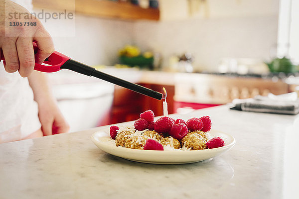 Ein Teller mit einem Dessert und frischen Himbeeren und einer Hand  die ein Feuerzeug für die Kerze hält.