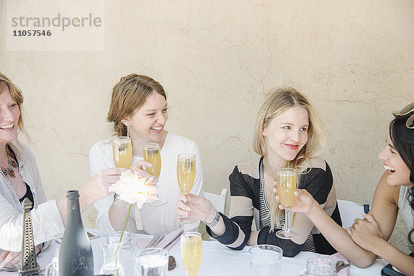 Vier lächelnde Frauen sitzen an einem Tisch  halten Gläser mit Champagner und stoßen an.
