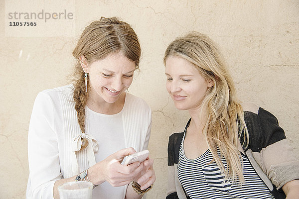 Zwei lächelnde Frauen mit langen blonden Haaren sitzen an einem Tisch und schauen auf ein Mobiltelefon.