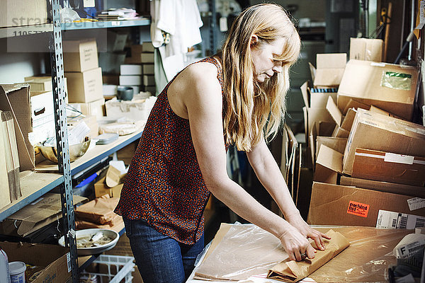 Frau mit langen blonden Haaren steht im Lagerraum eines Ladens und verpackt Waren in braunes Papier.
