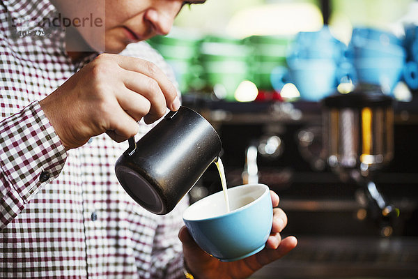 Spezialisiertes Kaffeehaus. Ein Mann gießt heiße Milch in eine Tasse Kaffee  um ein Muster auf der Oberseite zu machen.