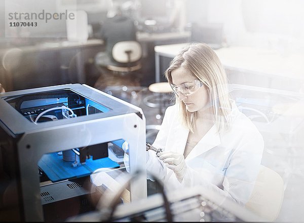 Technikerin  Frau 25-30 Jahre  mit weißem Laborkittel und Schutzbrille kontrolliert Druckvorgang von einem 3D Drucker  Wattens  Innsbrucker Land  Tirol  Österreich  Europa