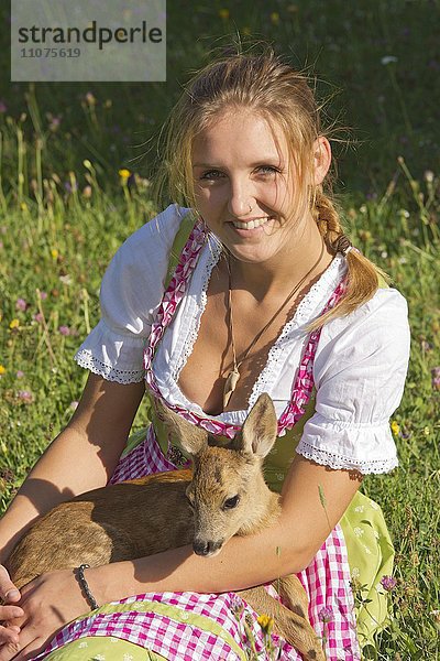 Frau im Dirndl mit einem zahmen Rehkitz auf dem Schoß sitzt in einer Blumenwiese  Tirol  Österreich  Europa