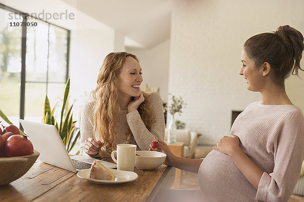 Schwangere Frauen essen Kuchen und Obst am Laptop im Esszimmer