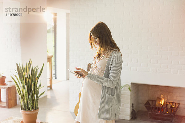 Schwangere Frau mit digitalem Tablett am Kamin im Wohnzimmer