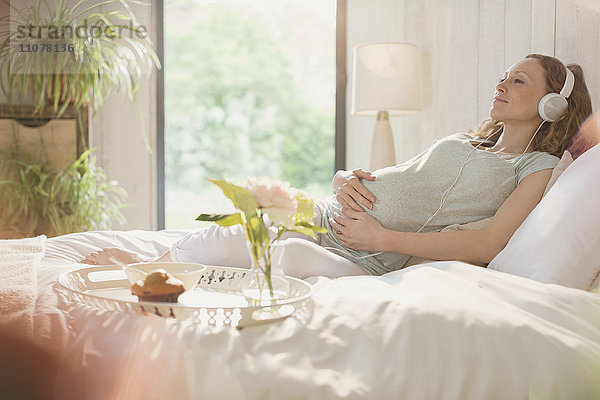 Gelassene Schwangere beim Musikhören mit Kopfhörern im Bett