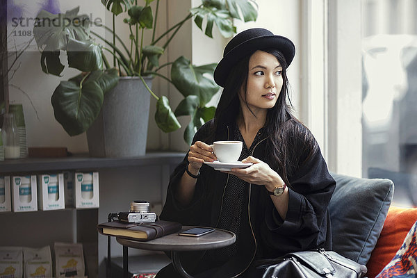 Nachdenkliche junge Frau mit Kaffeetasse im kreativen Büro am Fenster sitzend