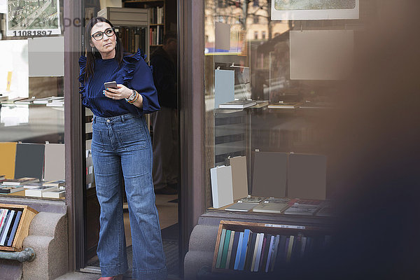 Nachdenkliche Frau hält Telefon  während sie am Eingang des Buchladens steht.