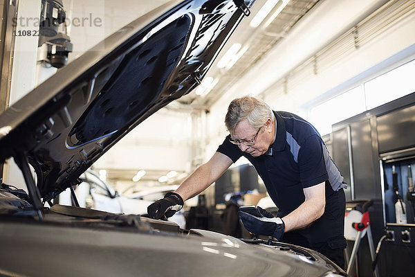 Senior Mechaniker hält Handy während der Reparatur des Autos in der Werkstatt