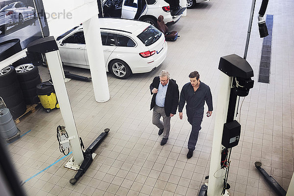 Hochwinkelansicht eines älteren Mannes  der mit einem Mechaniker während eines Spaziergangs in einer Autowerkstatt diskutiert.