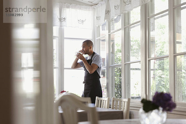 Männlicher Besitzer beim Kaffeetrinken und Telefonieren  während er am Fenster steht.