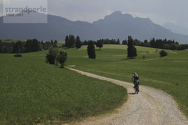 Rückansicht der Person  die mit dem Fahrrad auf einer unbefestigten Straße inmitten eines grasbewachsenen Feldes gegen die Berge fährt.