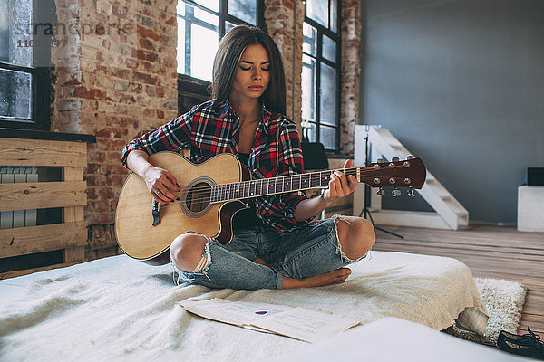 Junge Frau spielt Gitarre  während sie zu Hause auf dem Bett sitzt.